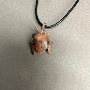 May include: A copper-colored pendant in the shape of a beetle hangs from a black cord necklace. The beetle pendant has detailed legs, antennae, and a textured surface. The necklace is displayed against a gray background.