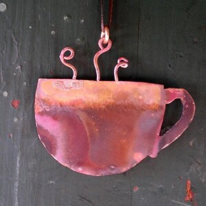 May include: A copper-coloured coffee cup ornament with a handle and steam rising from the top. The cup has a rustic, aged appearance.