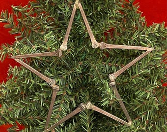 Puede incluir: Un adorno de Navidad plateado en forma de estrella hecho de clavos. El adorno está colgado de un árbol de Navidad artificial verde.