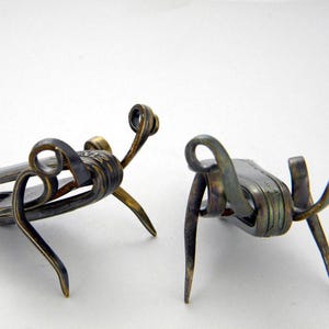 May include: Two small, silver-colored metal sculptures of insects, possibly grasshoppers, with intricate details. The sculptures are made from recycled materials, such as spoons or forks.