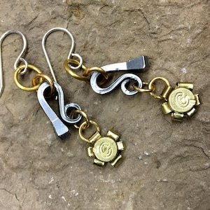 May include: Handmade earrings with a distinctive design. The earrings combine silver-tone metal hooks and charms with gold-tone accents. Each earring has a small, gold-tone charm with a circular design. The earrings are displayed on a textured, stone-like surface.