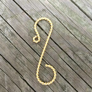 May include: A gold-colored, twisted metal ornament in an S-shape. The ornament is made of a braided material, with a loop at the top and a spiral at the bottom. The background is a weathered wooden surface.