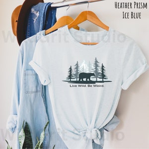 May include: A light blue t-shirt with a graphic of a bear walking through a forest with the text "Live Wild. Be Weird."