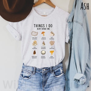 May include: A white t-shirt with the text "Things I do in my spare time" and a list of mushroom-related activities, including "Think about mushrooms", "Look for mushrooms", "Find mushrooms", "Cook mushrooms", "Talk about mushrooms", "Eat mushrooms", "Dream about mushrooms", "Research mushrooms", and "Hoard mushrooms". Each activity is illustrated with a different type of mushroom.