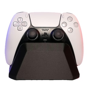 Könnte beinhalten: Ein weißer PlayStation 5 DualSense Wireless-Controller mit schwarzen Tasten und einem schwarzen Ladeständer.