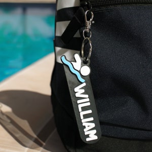 Pode incluir: Mochila preta com padrão listrado branco e preto. Um chaveiro personalizado em preto e branco com um ícone de nadador e o nome "WILLIAM" está preso ao zíper.