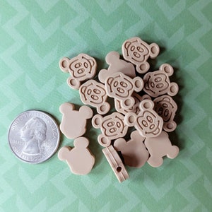 Könnte beinhalten: Ein Haufen tanfarbener Plastikknöpfe in Form von Mäuseköpfen mit einem schwarzen Mickey-Mouse-Gesichtsdesign. Die Knöpfe sind etwa so groß wie eine deutsche 2-Euro-Münze.