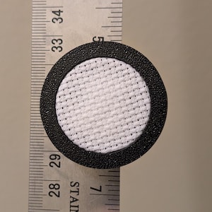 Pode incluir: Um filtro redondo preto com um centro de malha branco, com aproximadamente 3,2 cm de diâmetro, está posicionado ao lado de uma régua de aço inoxidável. A borda preta texturizada do filtro contrasta com a malha branca fina.