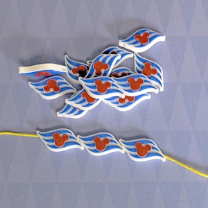 以下が含まれることがあります： 青と赤のミッキーマウスのデザインが施された白いプラスチック製のチャームのセット。チャームは、中央にミッキーマウスの頭が付いた波の形をしています。チャームは黄色の紐に通されています。