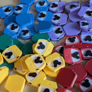 May include: A collection of colorful, hexagonal-shaped buttons with a Mickey Mouse silhouette design. The buttons are in various colors, including blue, green, yellow, red, and purple. The buttons are scattered on a light-colored surface.