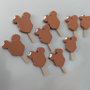 May include: Nine brown Mickey Mouse shaped ice lollies on wooden sticks. The lollies have a white bite taken out of them.