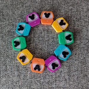May include: A circular arrangement of ten small plastic containers in various colors including purple, orange, yellow, green, and blue. Each container features a white lid with a black silhouette of a mouse head shape centered on the top surface.