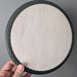 May include: A circular embroidery hoop frame featuring a natural beige linen fabric stretched tightly across the center. The outer edge of the hoop is finished with a dark forest green braided trim that provides a textured border around the fabric.