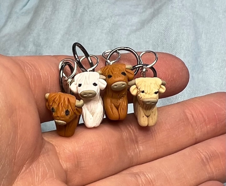 Op de afbeelding: Vier miniatuur Highland koe steekmarkeerders, elk met een andere kleur: bruin, wit, bruin en cr&egrave;me. De steekmarkeerders zijn bevestigd aan zilveren ringen.