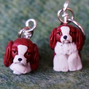 Könnte beinhalten: Zwei kleine, rote und weiße Cavalier King Charles Spaniel Hundefiguren aus Ton mit silbernen Sprungringen.