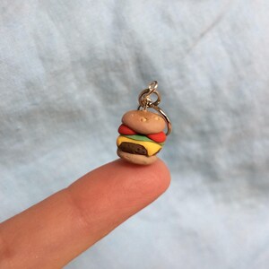 May include: A miniature clay hamburger charm with a brown bun, yellow cheese, green lettuce, and red tomato. The charm is attached to a silver key ring.