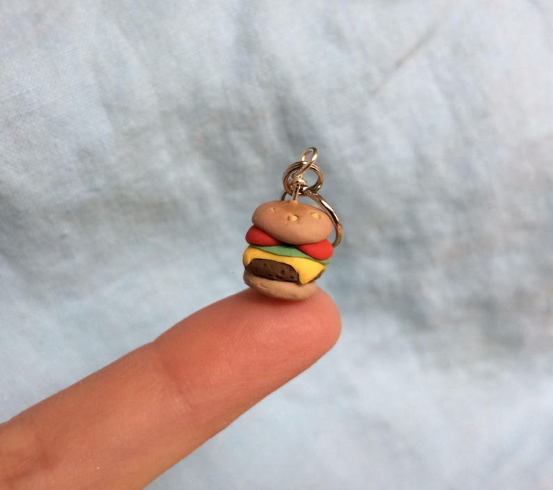 May include: A miniature clay hamburger charm with a brown bun, yellow cheese, green lettuce, and red tomato. The charm is attached to a silver key ring.