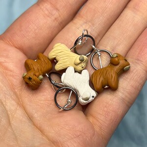 Op de afbeelding: Vier kleine, hondvormige steekmarkeerders in bruin, wit en beige. Elke markeerder heeft een zilveren ring met een zwarte draad.