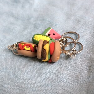 May include: A keychain with a variety of food charms, including a watermelon, corn on the cob, hot dog, and hamburger. The charms are attached to a silver key ring.