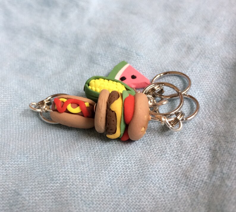 May include: A keychain with a variety of food charms, including a watermelon, corn on the cob, hot dog, and hamburger. The charms are attached to a silver key ring.