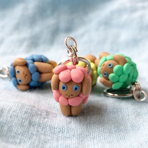 May include: Three small sheep charms made of polymer clay. The sheep are pink, blue, and green. Each sheep has a brown face with blue eyes and a silver loop for hanging.