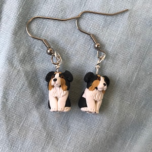 Può includere: Un paio di orecchini fatti a mano con figurine di cani in miniatura. Ogni cane ha uno schema di colori nero, bianco e marrone, con ganci argentati. Gli orecchini sono su uno sfondo di tessuto azzurro.