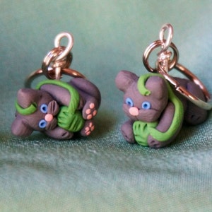 May include: Two gray cat keychains with green yarn, blue eyes, and pink noses. The keychains are made of polymer clay and have silver key rings.