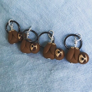May include: Four brown sloth-shaped stitch markers with black rings and silver hooks. The sloths are hanging upside down.
