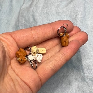 Op de afbeelding: Vier kleine, handgemaakte, dierlijke steekmarkeerders in de vorm van Highland-koeien. De steekmarkeerders zijn bruin, wit en beige. Ze zijn bevestigd aan zilveren ringen.