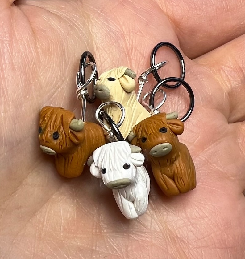 Op de afbeelding: Vier handgemaakte polymer clay koe-steekmarkeerders met zilveren ringen. De koeien zijn bruin, wit en cr&egrave;me.