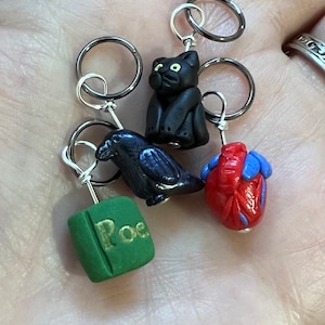 Op de afbeelding: Set van vijf zwarte en zilveren steekmarkeerders met verschillende bedels. De bedels omvatten een zwarte kat, een raaf, een rood hart en een groen boek met het woord "Poe" erop.
