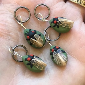 May include: Four green and brown cicada stitch markers with silver rings. The cicadas have black bodies, red eyes, and translucent wings.