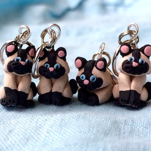 May include: Four small, handmade, polymer clay Siamese cat charms with silver key rings. The cats are sitting with their paws tucked under them and have black and white markings with blue eyes.