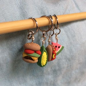 May include: Four colorful food-themed stitch markers, including a hamburger, corn on the cob, hot dog, and watermelon slice, are hanging on a wooden dowel.