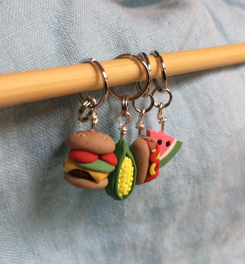 May include: Four colorful food-themed stitch markers, including a hamburger, corn on the cob, hot dog, and watermelon slice, are hanging on a wooden dowel.