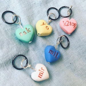 May include: Five heart-shaped stitch markers with different knitting abbreviations. The markers are made of clay and have black and silver rings attached. The abbreviations are "ssk", "yo", "k2tog", "mil", and "mir".
