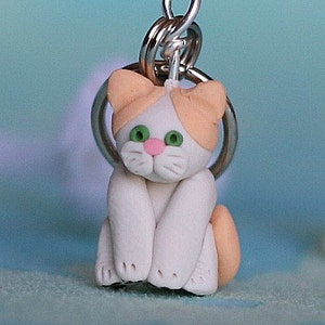 May include: A white and peach-colored cat charm with green eyes, a pink nose, and a silver key ring. The cat is sitting with its paws together and its tail curled around its body.
