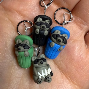 May include: Four handmade raccoon charms in various colors: green, black, blue, and gray. Each raccoon is sculpted with a trash can design and attached to a silver-tone keyring. The gray raccoon holds a bone-shaped object.