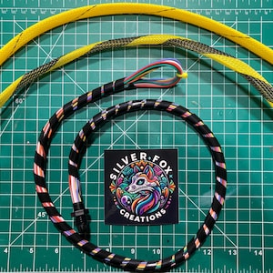 Puede incluir: Primer plano de un cable trenzado de colores con rayas negras, blancas, rosas y azules. El cable está envuelto en un tubo termocontraíble rayado negro y amarillo. El cable también está envuelto en una manga trenzada rayada amarilla y negra. El cable está enrollado en un círculo y está sobre una estera de corte verde. El texto "SILVER FOX CREATIONS" es visible en una pegatina.