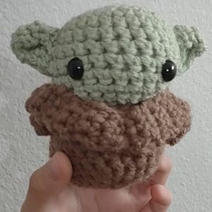 May include: A handmade crochet doll in the likeness of a popular character. The doll features a light green head with large black eyes and pointed ears, atop a brown body. The doll is held in a hand against a white background.