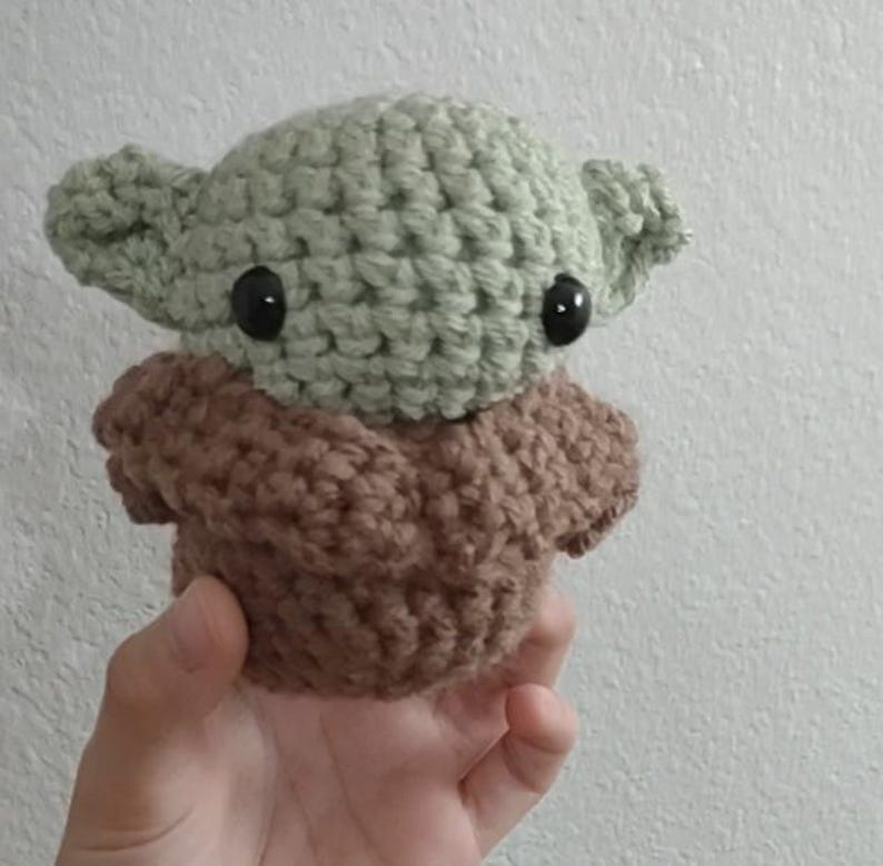 May include: A handmade crochet doll in the likeness of a popular character. The doll features a light green head with large black eyes and pointed ears, atop a brown body. The doll is held in a hand against a white background.