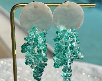 Könnte beinhalten: Ohrringe zum Hängen mit einem runden, perlmuttartigen Oberteil und einer Kaskade aus kleinen, hellgrünen Perlen. Die Ohrringe werden auf einem goldfarbenen Ständer vor einem verschwommenen Wasserhintergrund präsentiert.
