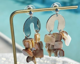 Könnte beinhalten: Silberfarbene Ohrringe mit gebogenem Oberteil und herabhängenden, gestapelten, rechteckigen Muschelteilen in Braun-, Beige- und Weißtönen. Die Ohrringe werden auf einem goldfarbenen Ständer vor einem verschwommenen blauen Hintergrund präsentiert.