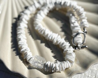 Peut inclure: Un collier blanc avec un pendentif en cristal transparent et un fermoir argenté. Le collier est composé de petits morceaux blancs irréguliers. Le collier est présenté sur une assiette blanche en forme de coquillage.