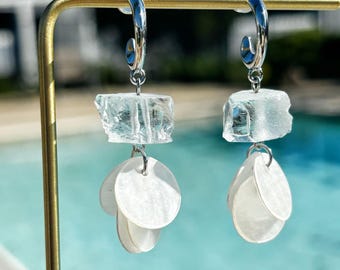 Könnte beinhalten: Silberne Creolen mit einem klaren, strukturierten, rechteckigen Stein und drei weißen, runden, muschelartigen Anhängern. Die Ohrringe werden vor einem verschwommenen Hintergrund aus einem Pool und einem goldfarbenen Metallständer präsentiert.
