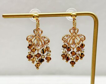 Könnte beinhalten: Ein Paar goldfarbene Chandelier-Ohrringe. Jeder Ohrring hat ein verziertes Filigran-Design mit herabhängenden Clustern aus braunen und klaren Perlen. Die Ohrringe werden auf einem goldenen Ständer vor weißem Hintergrund präsentiert.