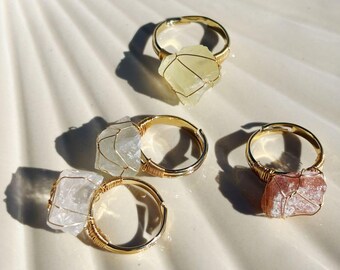 Könnte beinhalten: Vier goldfarbene Ringe mit natürlichen Steineinfassungen. Die Steine sind in verschiedenen Farben, darunter klar, gelb und rosa, und in Draht gewickelt. Die Ringe werden auf einer weißen, strukturierten Oberfläche präsentiert.
