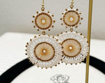 Könnte beinhalten: Handgefertigte weiße Perlenohrringe mit goldfarbenem Mittelteil und goldfarbenen Akzenten. Die Ohrringe haben ein kreisförmiges Design mit einem kleineren Kreis auf einem größeren. Die Ohrringe werden auf einem goldfarbenen Ohrringständer präsentiert.