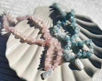 Könnte beinhalten: Zwei Halsketten mit Edelsteinperlen und Perlenanhängern liegen auf einer muschelförmigen Schale. Eine Halskette hat rosa Perlen, die andere blaue. Die Schale ist cremefarben, und der Hintergrund ist unscharf.