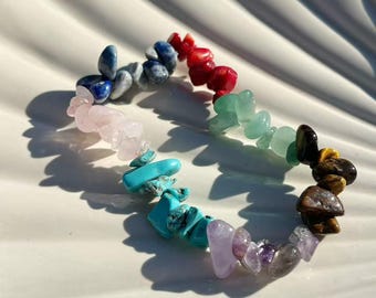 Könnte beinhalten: Ein farbenfrohes Edelsteinarmband mit polierten Steinen in Blau, Rot, Grün, Gelb, Lila und Rosa. Das Armband liegt auf einem weißen, muschelförmigen Teller, der die natürliche Schönheit der Steine hervorhebt.